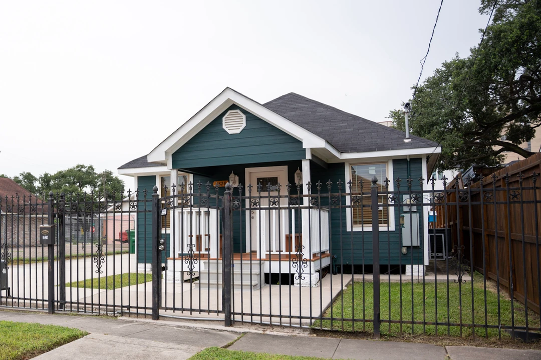 A blue bungalow with a white porch behind an iron fence.