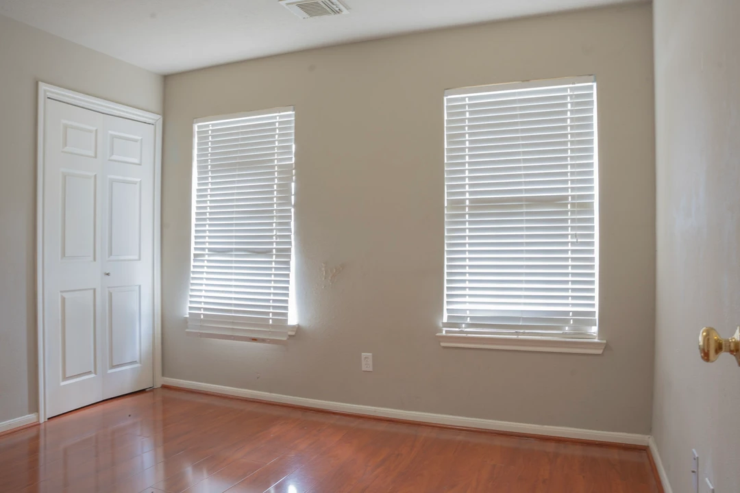 An empty bedroom with wood flooring and two windows with white blinds.
