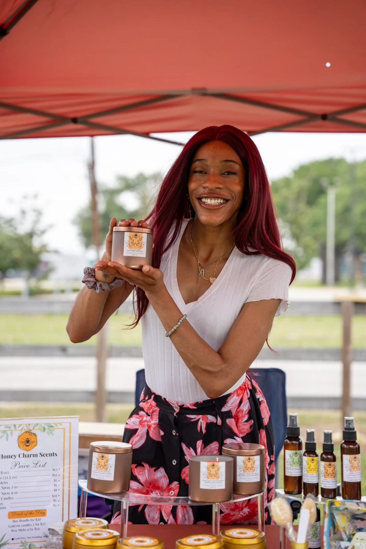 A smiling candle maker holds a jar beside her Honey Charm Scents display.