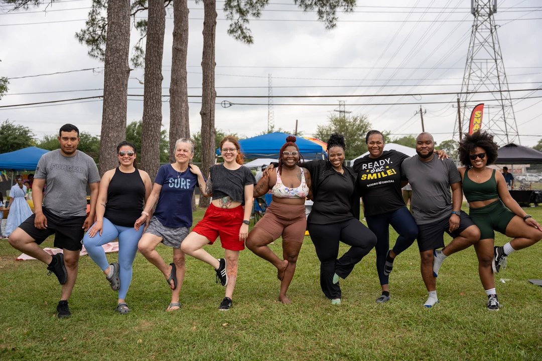 Market visitors balance in tree pose together on a green lawn.
