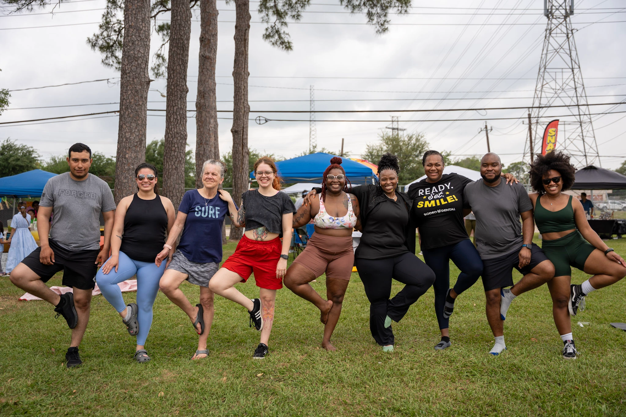 Market visitors balance in tree pose together on a green lawn.