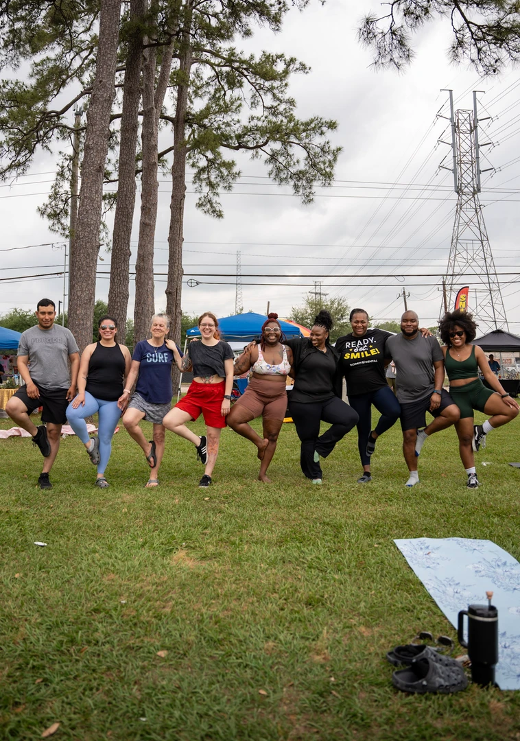 Market visitors balancing in tree pose together on the lawn.