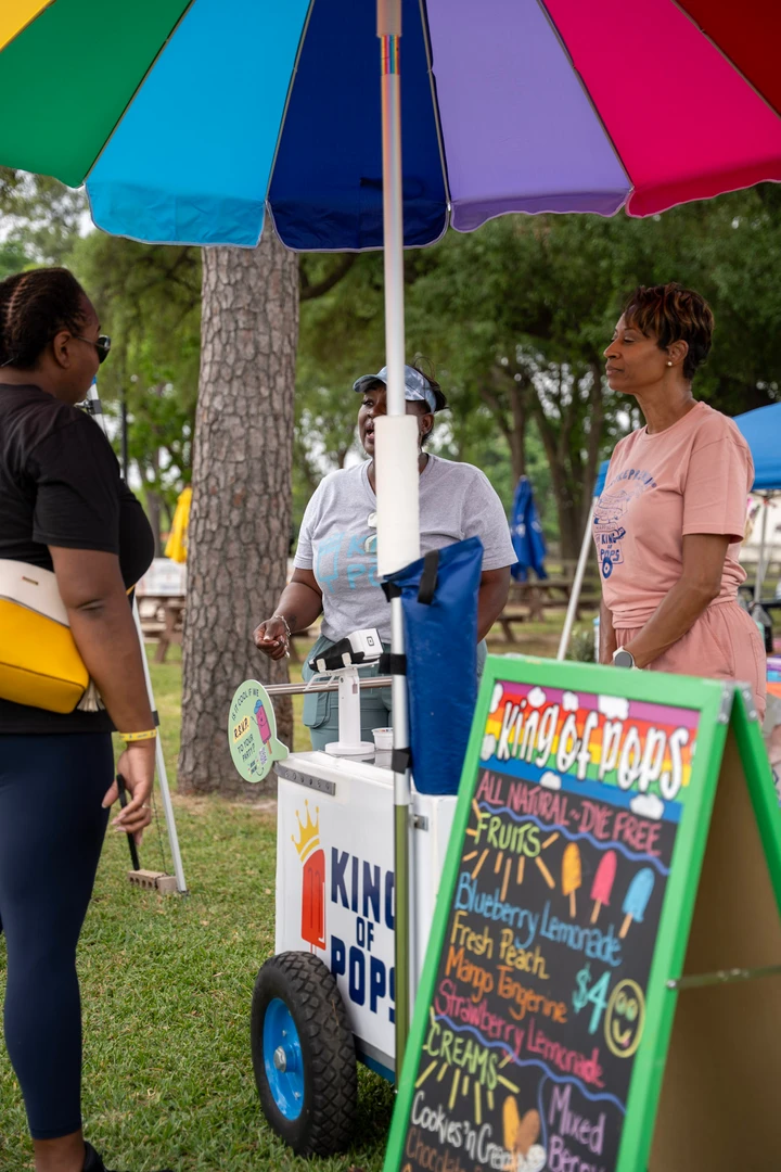 Visitors chatting at a drink cart beneath a rainbow umbrella.