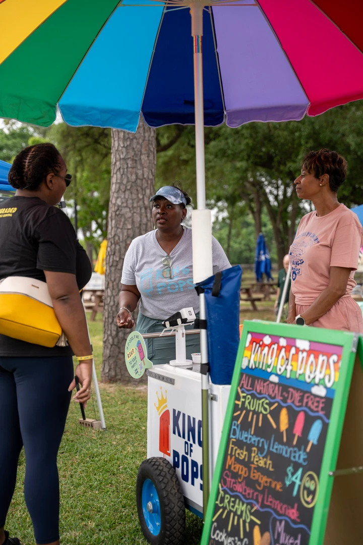 Visitors chatting at a drink cart beneath a rainbow umbrella.