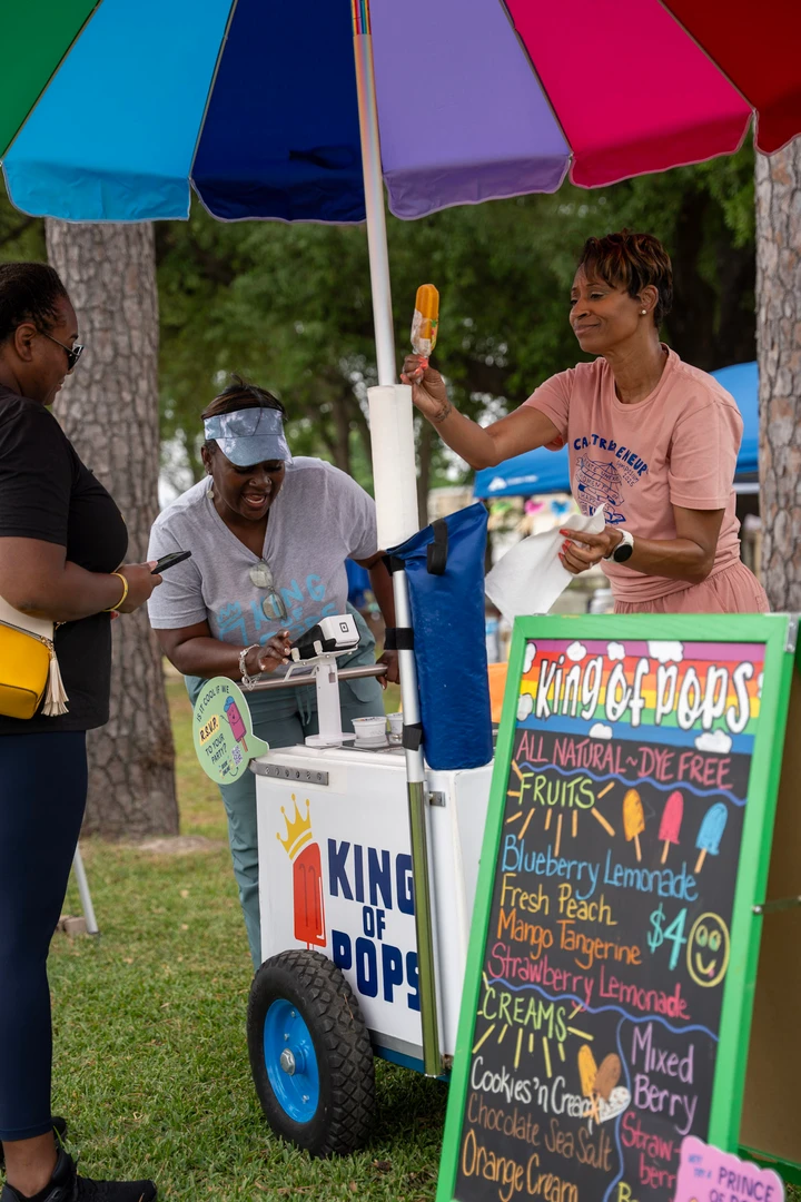 Visitors chatting at a drink cart beneath a rainbow umbrella.