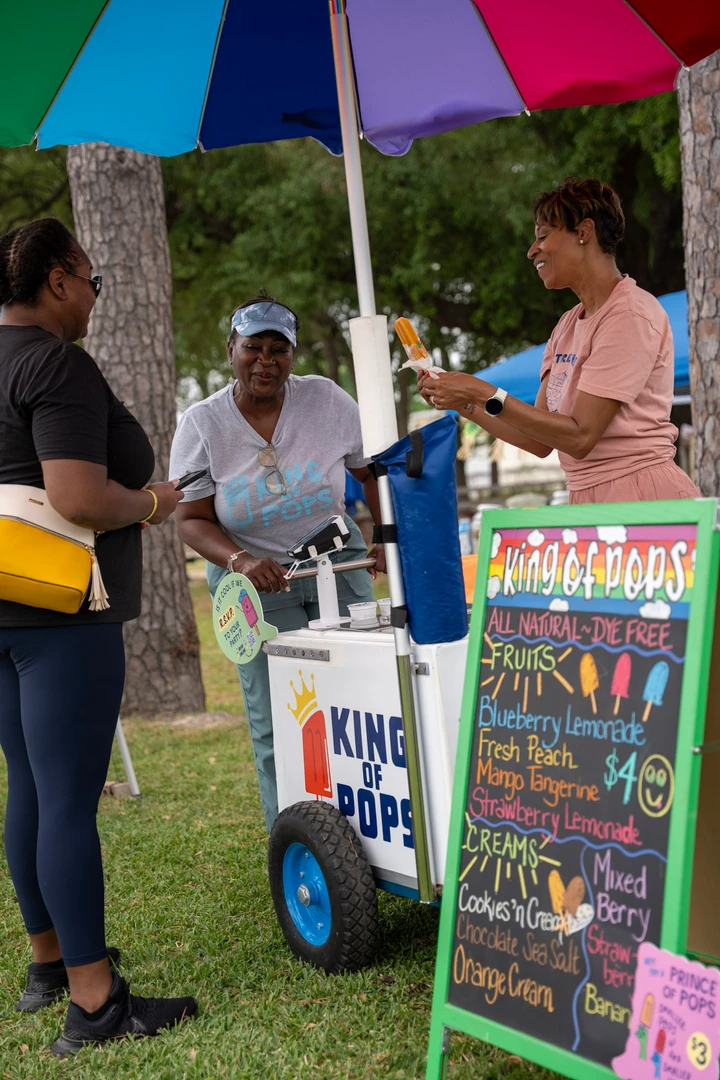 Visitors chatting at a drink cart beneath a rainbow umbrella.