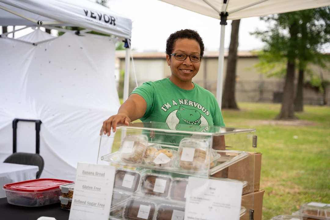 A smiling baker in green behind a clear display of baked goods.