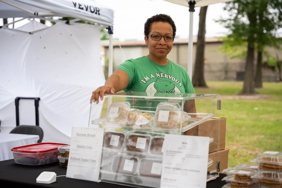 A smiling baker in green behind a clear display of baked goods.