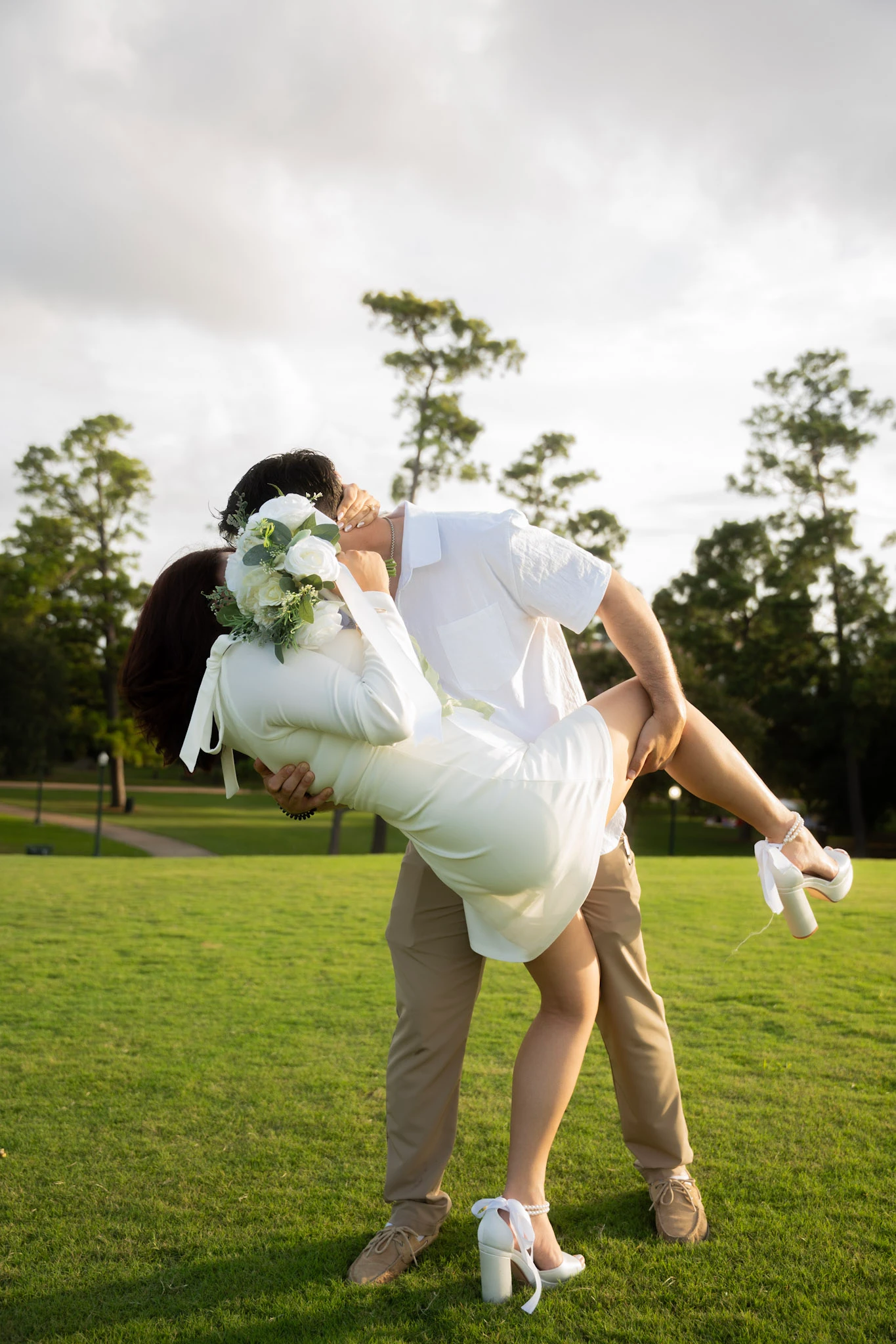 A couple share a kiss during a playful dip in a green park.