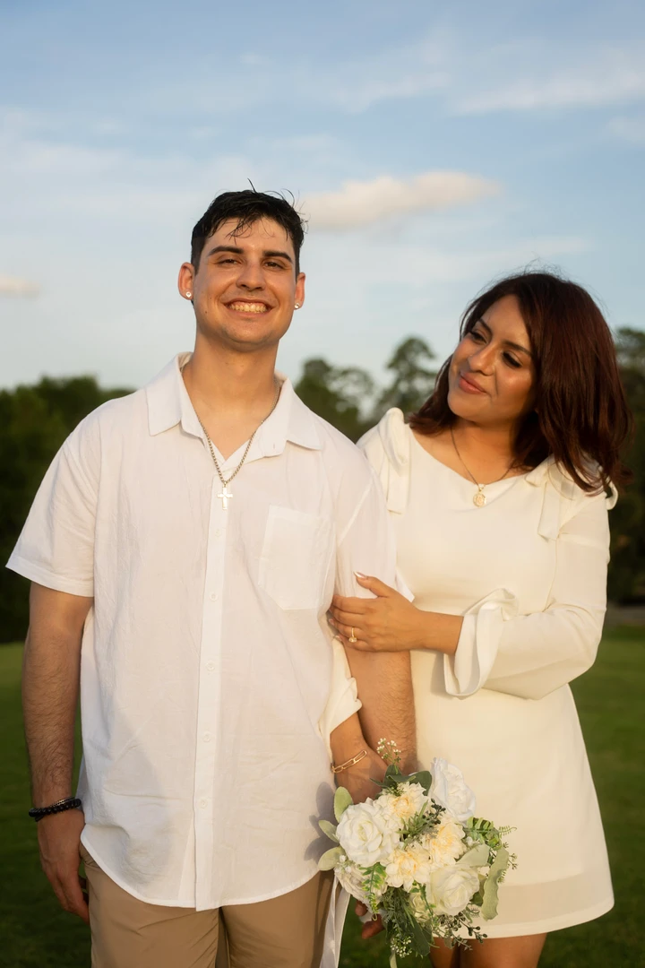 A couple in white hold hands and a bouquet in soft evening light.