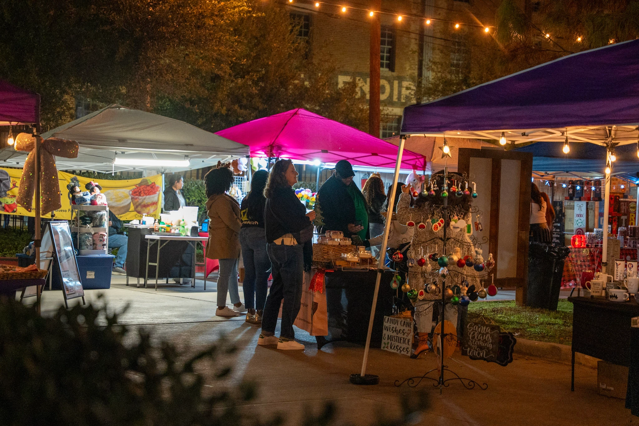 Holiday market tents and shoppers glow under strings of warm lights.