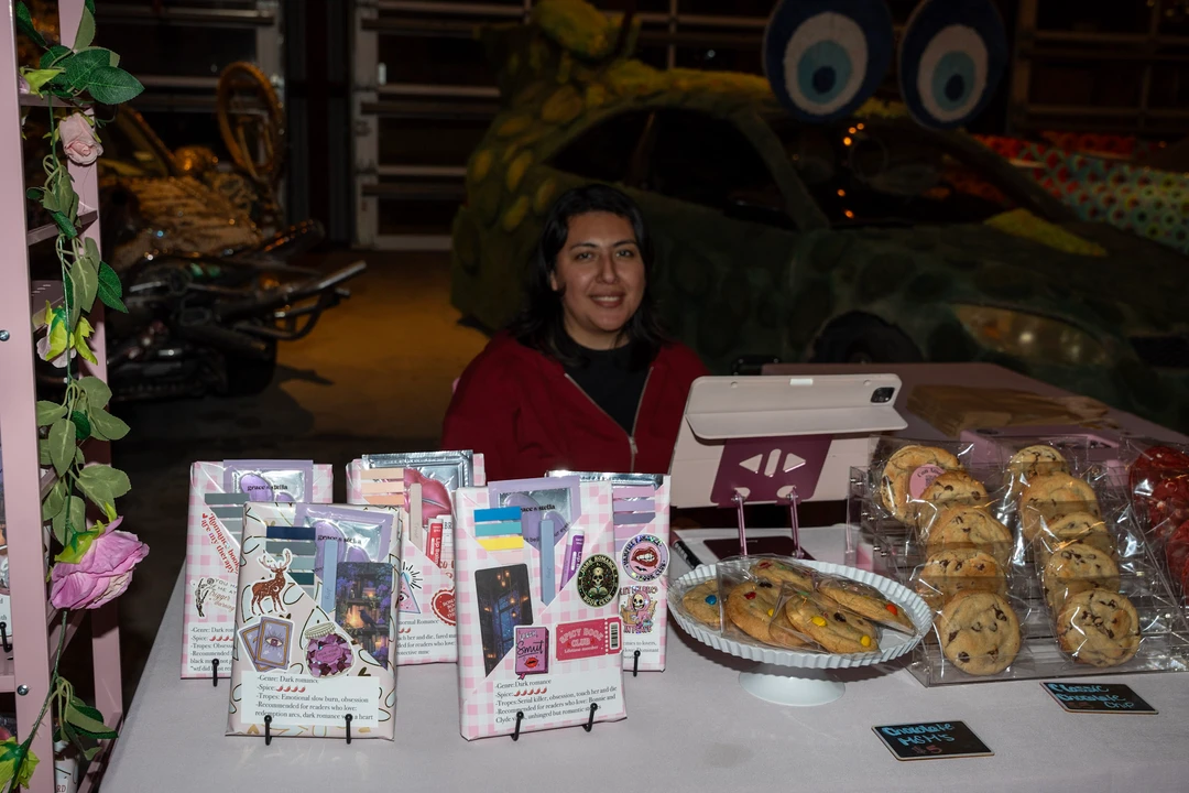 A smiling vendor seated behind handmade gifts and baked goods.