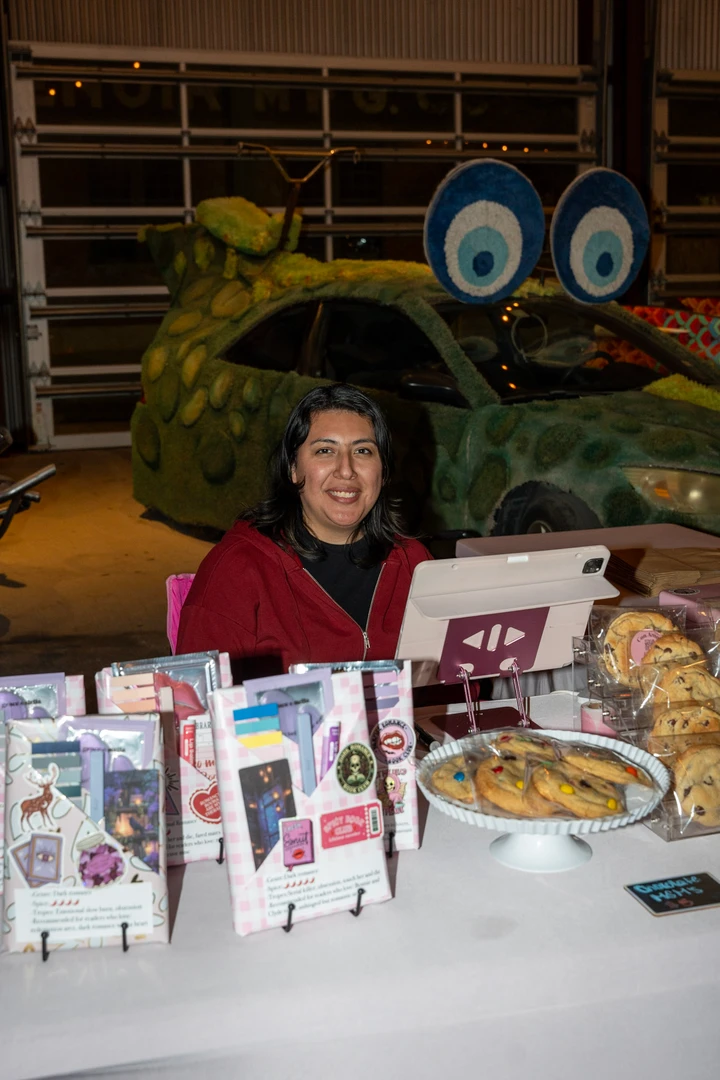 A smiling vendor seated behind handmade gifts and baked goods.