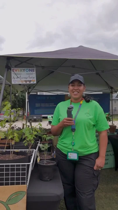 A plant vendor in a green shirt speaking beside potted plants at a market.